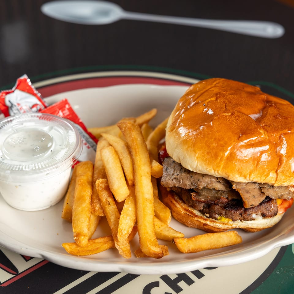 Double-patty burger with a glossy brioche bun, fries, dipping sauce, and condiment packets on a diner plate.